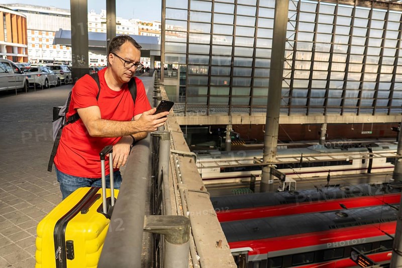 Solo male traveler waiting at a modern railway station, holding a yellow suitcase and checking his phone while looking over the train platforms below