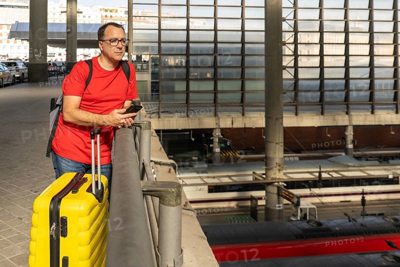 Adult male traveler waiting at an elevated railway station, holding a yellow suitcase while observing trains below in a modern urban transportation setting