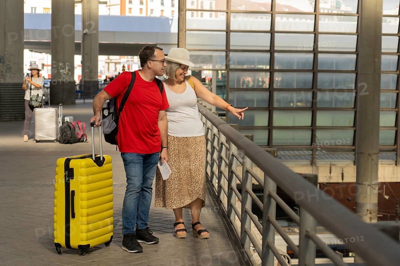 Adult travel couple walking together inside a modern transportation terminal above train tracks, pulling luggage while talking and pointing. Multimodal transport, travel connection, tourism and urban mobility concept