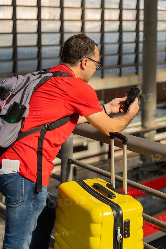 Mature male traveler using a smartphone at a train station while standing next to a yellow suitcase. Concept of travel, technology, urban mobility and modern tourism