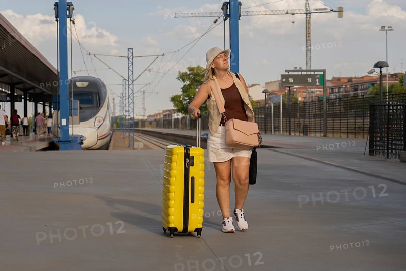 Adult woman walking along a modern train station platform, pulling a yellow suitcase while waiting for her train during a relaxed urban travel moment