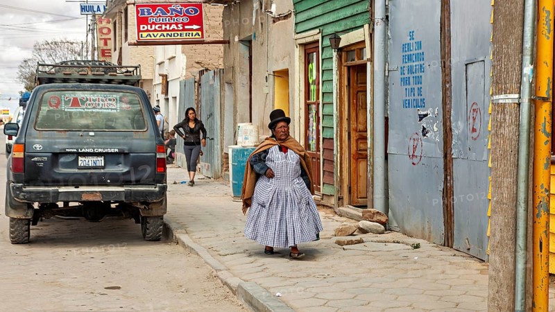 Woman wearing traditional clothes wearing melon walks on a sidewalk with colorful houses and murals, people in an altiplano near Uyuni in Bolivia