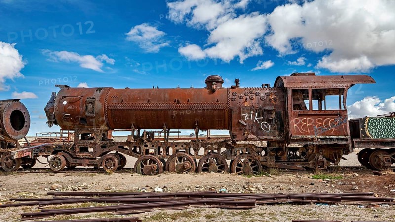 Rusted locomotive under a cloudy blue sky with graffiti along the body, old rusty locomotives at the train cemetery near Uyuni in the Altiplano in Bolivia