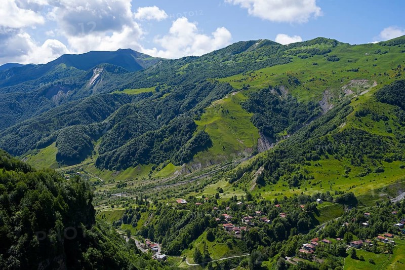 Lush green mountains with scattered village under blue sky and clouds, viewpoint near Gudauri, view of the mountainous landscape around Mleta, Mtskheta-Mtianeti region, Georgian Military Highway, High Caucasus, Georgia