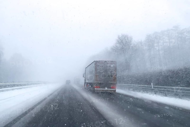 View through the windshield of a passenger car driving through light snowblowing on partly snow-covered motorway with lanes covered by snow, Germany