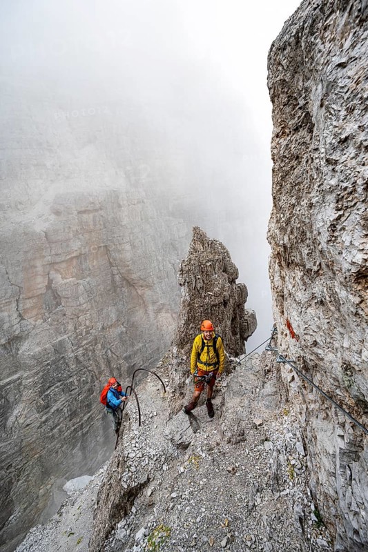 Two mountaineers climb the Via Ferrata Oliva Detassis via ferrata in fog, steep mountains covered in clouds, Brenta Mountains, Trentino, Italy