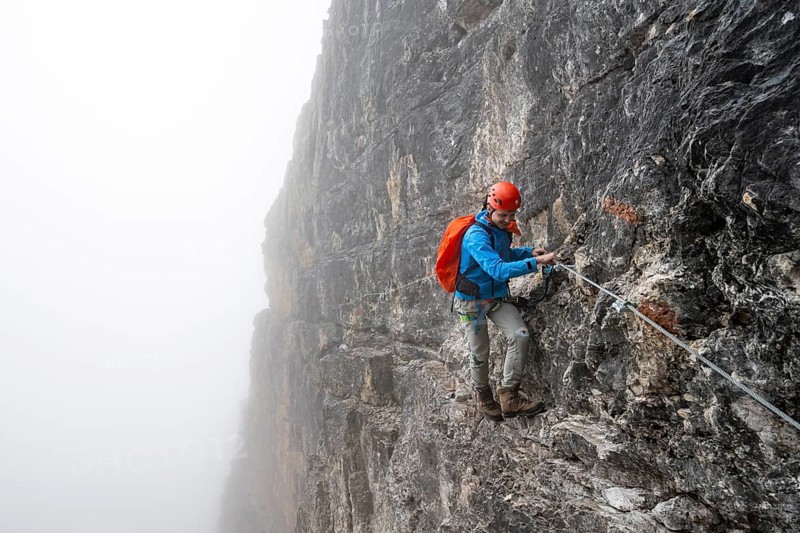 Mountaineers on a steep rock face on the Via Ferrata Oliva Detassis via ferrata in fog, steep mountains covered in clouds, Brenta Mountains, Trentino, Italy