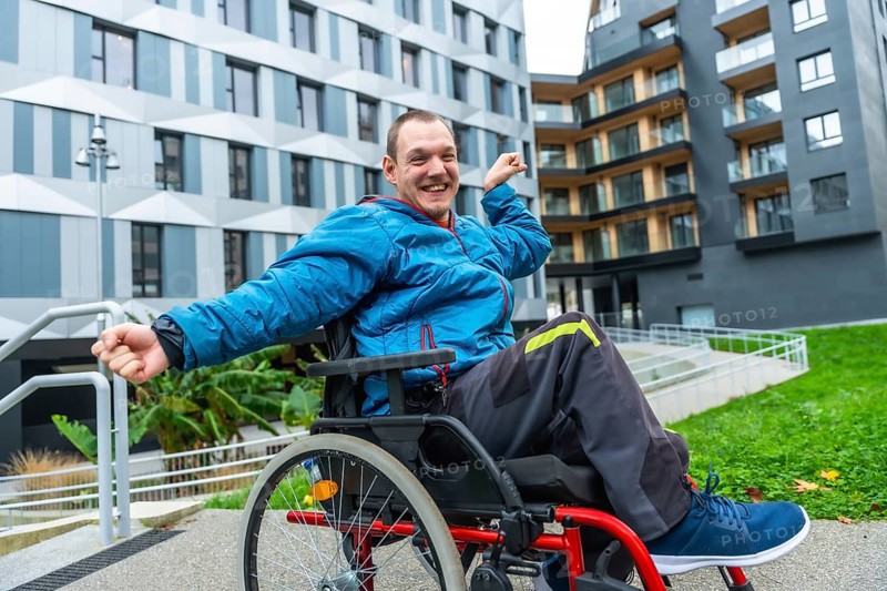 Man in wheelchair smiling and raising fists in victory on a city sidewalk by a modern building and accessible ramp, expressing independence, positivity and active urban living