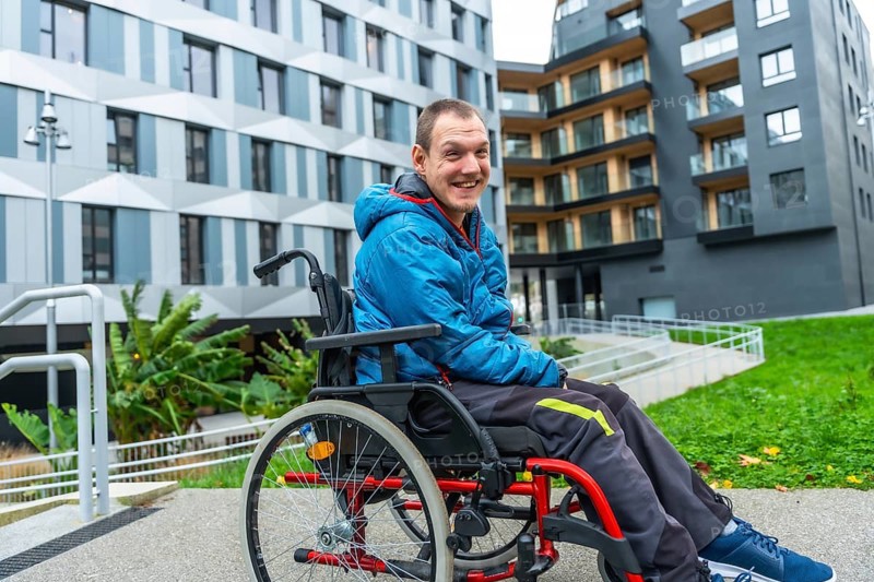 Happy man with a disability using a wheelchair on an accessible ramp in front of modern buildings, symbolizing inclusion, independent living, and urban accessibility for people with special needs