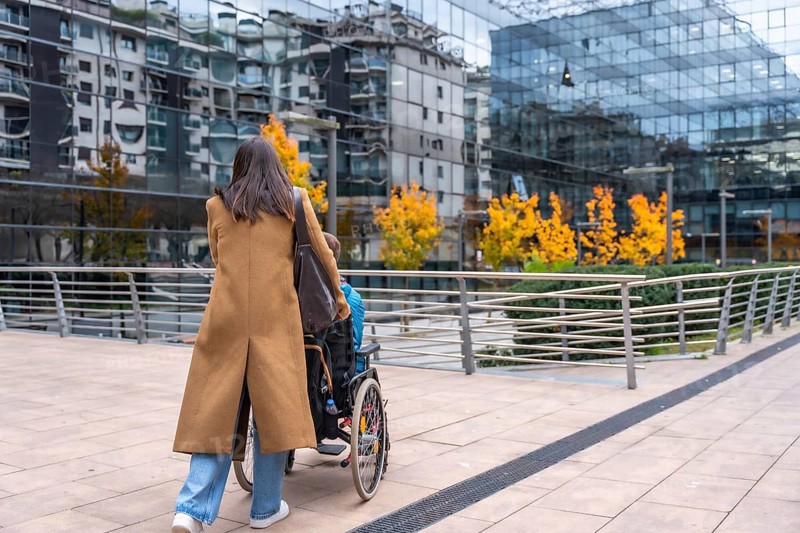 Woman in a brown coat pushing a young person in a wheelchair along a modern urban sidewalk, navigating the city street with glass buildings and autumn trees