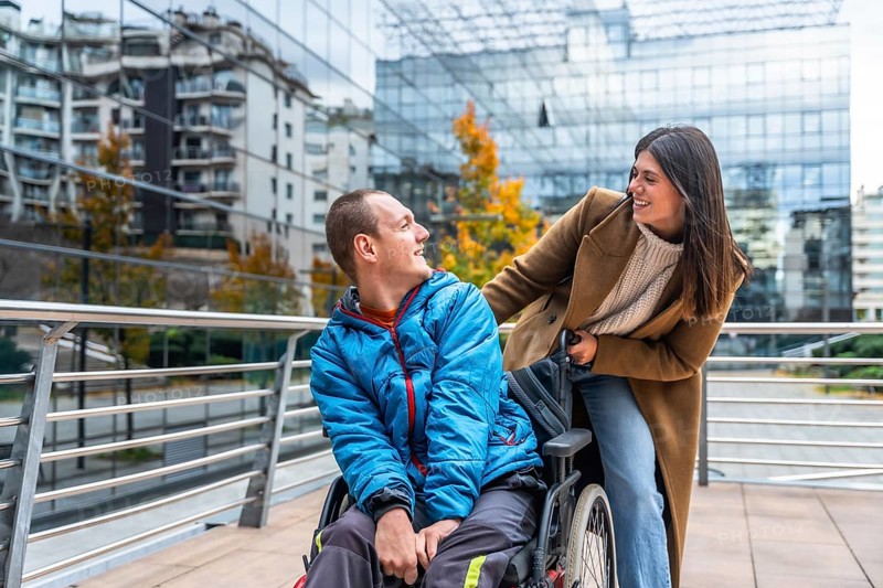 Caregiver woman pushes smiling young man in wheelchair along a city sidewalk, sharing a warm moment of support, inclusion and friendship amid urban architecture and trees