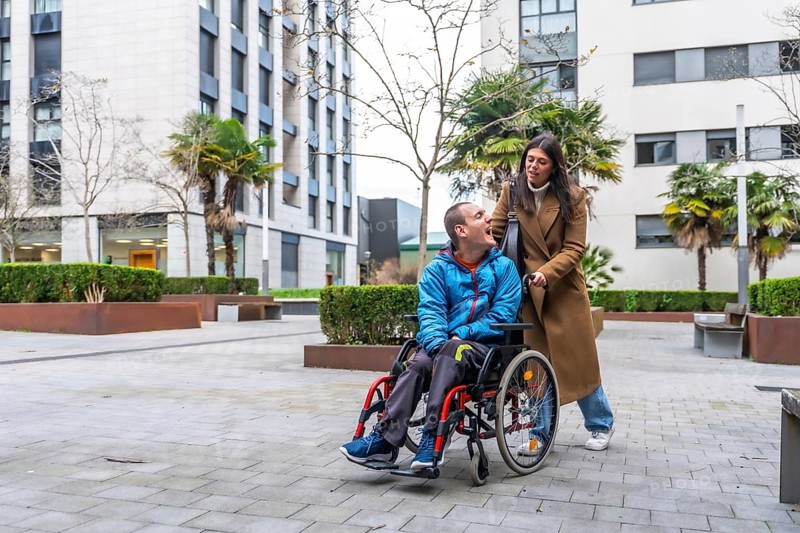 Woman pushing smiling man in a wheelchair along a city sidewalk, conveying everyday support, accessibility, companionship and inclusive, independent urban living