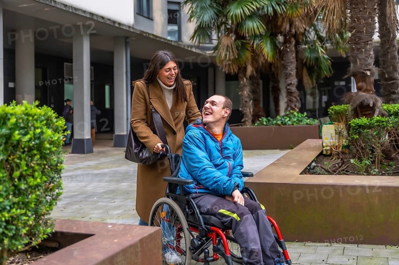 Smiling caregiver pushing wheelchair, sharing warm conversation and laughter with happy man outdoors, promoting inclusion, companionship and active assisted living in urban setting