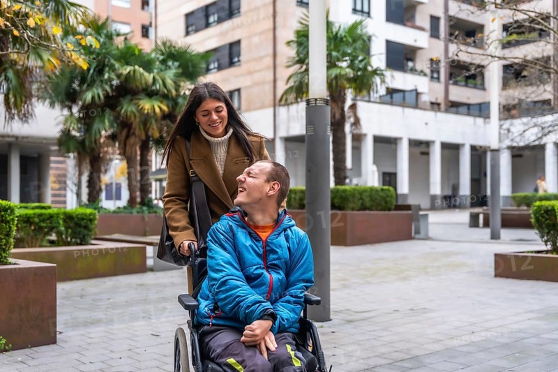 Young woman pushing a man in a wheelchair, both smiling while enjoying an outdoor stroll in an urban area, highlighting care, assistance, and inclusion