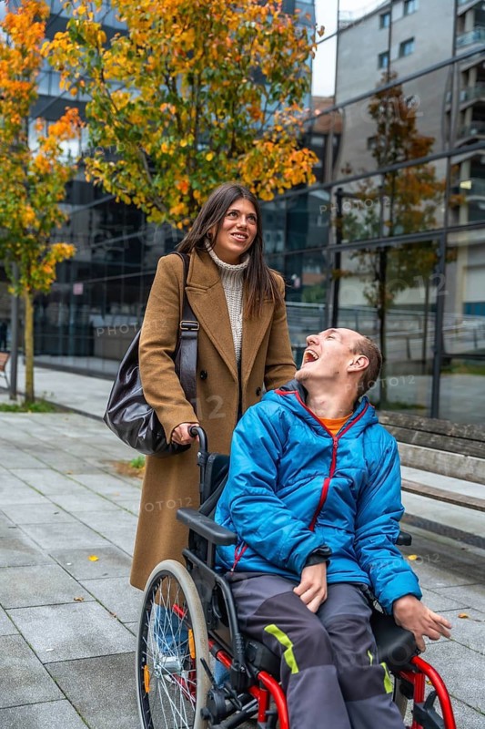 Woman pushes cheerful man in a wheelchair along a city sidewalk lined with autumn trees, sharing laughter and support during an active, inclusive outdoor moment