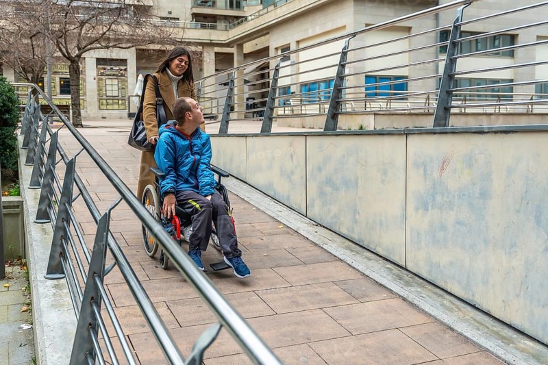 Woman pushing a young man with a disability in a wheelchair, navigating an accessible ramp with railings in an urban setting, receiving support and care