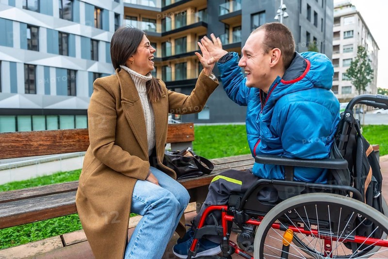 Woman and disabled man in wheelchair laughing, high fiving, and sharing a happy moment during an outdoor session, symbolizing support, friendship, and positive connection
