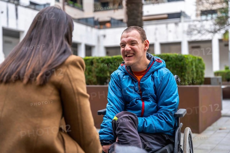 Young disabled man with a cheerful expression sitting in a wheelchair and engaging in a supportive conversation with a woman outdoors, highlighting inclusion and positive interaction