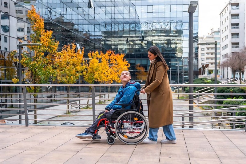 Woman pushing a smiling man in a wheelchair along an urban sidewalk during autumn, showcasing support, companionship, and accessibility in a modern city environment