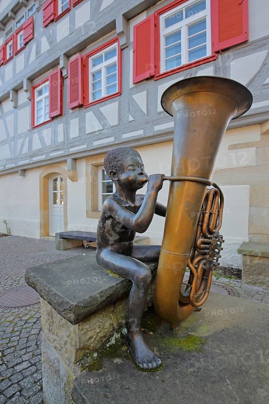 Sculpture boy with tuba by Guido Messer 1991, modern art, bronze sculpture, playing music, tuba, brass instrument, sitting, jack, size comparison, big, small, big cheeks, blowing, half-timbered house, music school, Waldenbuch, Baden-Württemberg, Germany