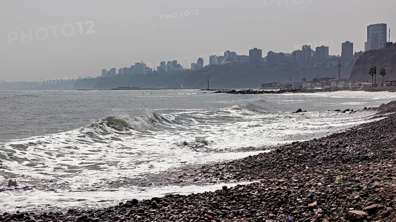 Pebble beach by the sea with waves, in the background skyscrapers of a city on a cloudy coastline, the coast with the skyline of Lima in Peru