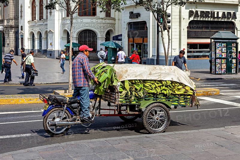 Motorcycle trailer loaded with bananas on a busy city street at an intersection, a man on a motorcycle with a load of bananas in downtown Lima in Peru