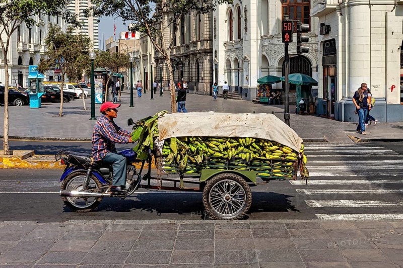 Person on motorcycle with trailer full of bananas in an urban setting at an intersection, a man on a motorcycle with a load of bananas in downtown Lima in Peru