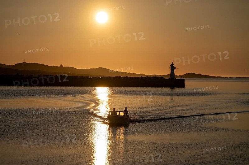 A small motorboat sails through the harbor entrance with lighthouse and midnight sun, Vardø, Finnmark, Norway