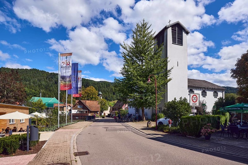 St. Peter and Paul parish church, church tower, flag on the flagpole, 750 years Gutach Schwarzwaldbahn, the home of the Bollenhut, ... always goes, general architecture, Black Forest house, hill, hilly landscape with coniferous forest, zone 30 sign, road, blue sky, cumulus clouds, Kirchstraße, Gutach Schwarzwaldbahn, Black Forest, Ortenaukreis, Baden-Württemberg, Germany