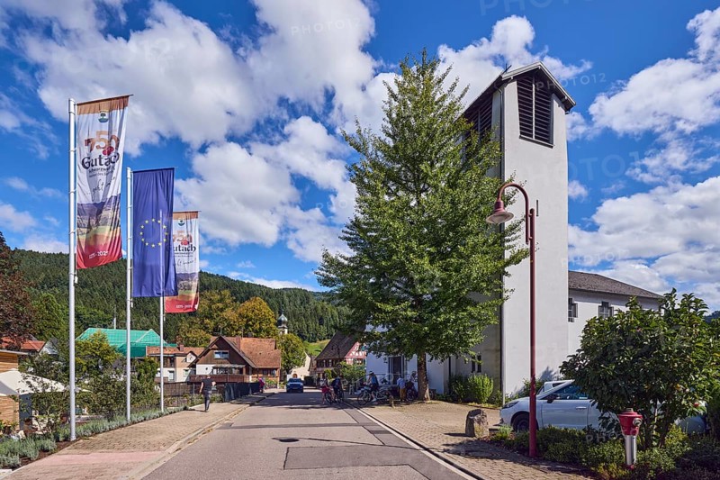 St. Peter and Paul parish church, church tower, flag on flagpole, European flag, 750 years Gutach Schwarzwaldbahn, the home of the Bollenhut, ... always goes, general architecture, Black Forest house, hill, hilly landscape with coniferous forest, road, pedestrians as accessories, blue sky, cumulus clouds, Kirchstraße, Gutach Schwarzwaldbahn, Black Forest, Ortenaukreis, Baden-Württemberg, Germany