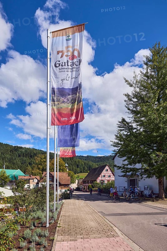 Flags on flagpoles, 750 years of Gutach Schwarzwaldbahn, The home of the Bollenhut, ... always goes, 1275 - 2025, sidewalk, concrete paving stones, hills, coniferous forest, trees, general architecture, Schwarzwaldhaus, blue sky, cumulus clouds, Kirchstraße, Gutach Schwarzwaldbahn, Black Forest, Ortenaukreis, Baden-Württemberg, Germany