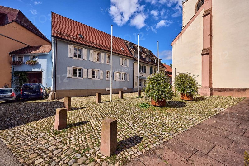 Houses, residential buildings, apartment blocks, church, facade with windows and shutters, dormers, flagpoles, planters with Cape Leadwort (Plumbago auriculata), bollards, cobblestone square, blue sky, cumulus clouds, Kirchstraße, Staufen im Breisgau, Black Forest, Breisgau-Hochschwarzwald district, Baden-Württemberg, Germany