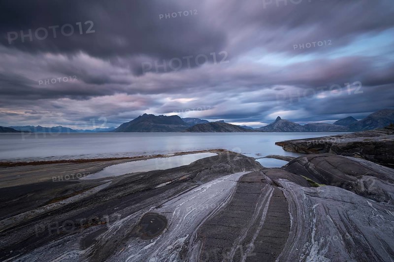 The Vestfjord in Nordland, Norway, offers a dramatic atmosphere near Bodø. Dark clouds are illuminated by the setting sun in shades of red and yellow. The rugged cliffs and mountains are reflected in the quiet fjord, which offers a breathtaking view of Lofoten