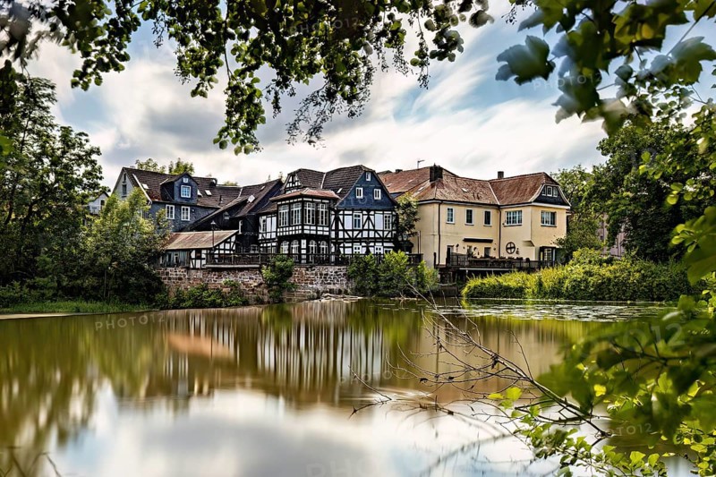 Half-timbered houses are reflected in the quiet lake, surrounded by lush vegetation. The historic houses of Marburg are reflected in the Lahn in Hesse