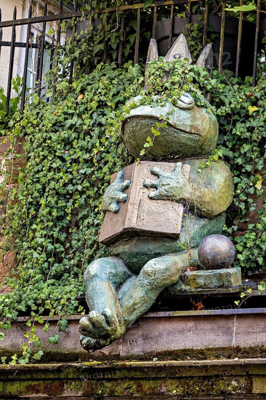 A frog sculpture with crown and book, surrounded by ivy, creates an enchanted atmosphere, The Frog King of Marburg an der Lahn in Hesse