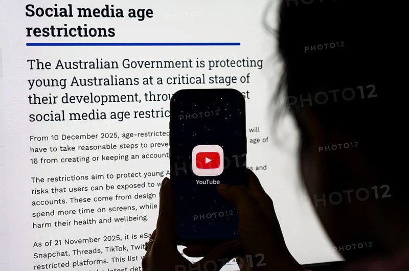 In this photo illustration, a person holds a phone displaying the YouTube app in front of a screen showing information about Australia's new social media age restrictions, in New Delhi, India on 12 December 2025