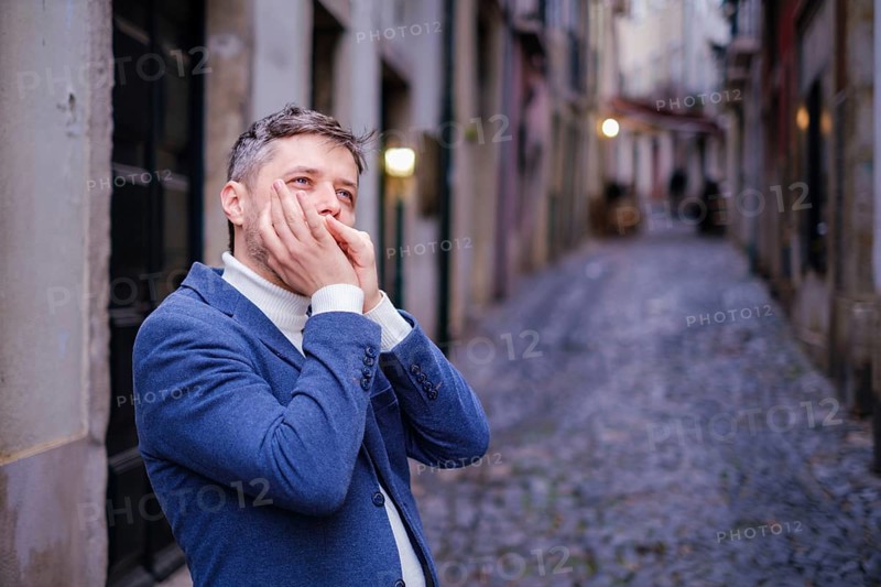 Man musician in a blue blazer and white sweater playing blues on a harmonica with eyes closed, standing outdoors in city street