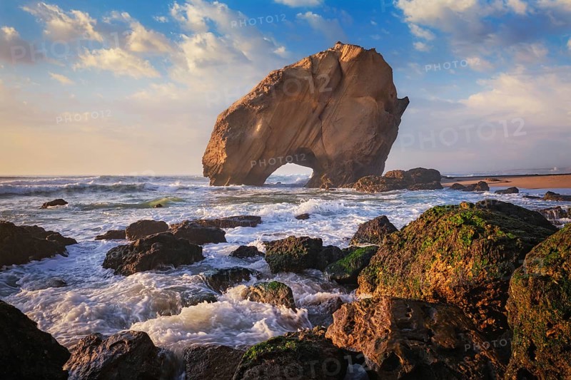 Penedo do Guincho, a large boulder rock arch at Praia da Santa Cruz, Portugal, with ocean waves and sandy beach on sunset