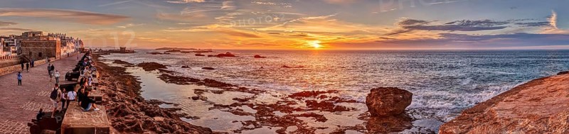 Panorama of a coastal promenade at sunset with rocks in the sea and a bright orange sky, Morocco, Essaouira