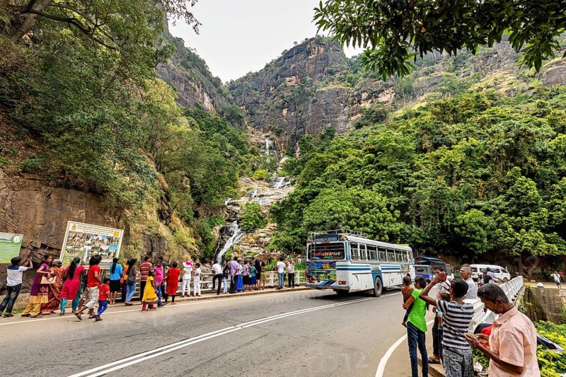 Mountain road with a bus and people looking at a waterfall surrounded by green mountain scenery, tourists at waterfalls in the mountains near Kandy in Sri Lanka