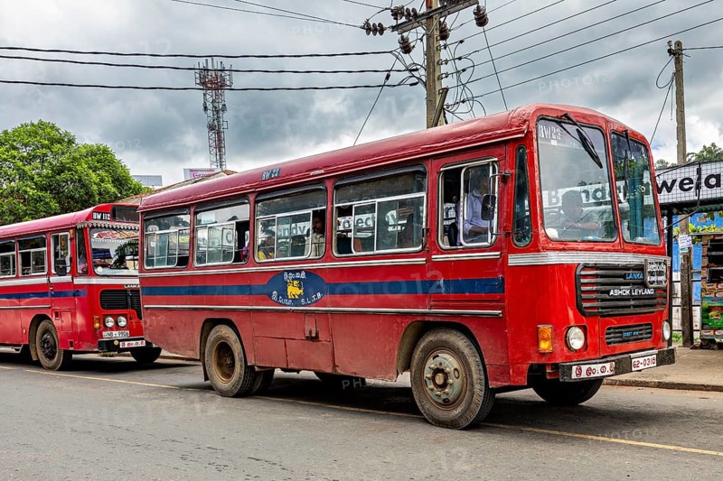 A red bus on a busy street with cloudy skies and power lines, The red buses in Sri Lanka