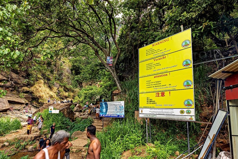 Trail in the jungle with warning signs and people surrounded by trees and rocks, tourists at the waterfalls in the mountains near Kandy in Sri Lanka