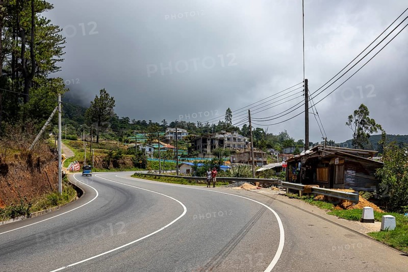 A winding road in a rural area with buildings, trees and a cloudy sky, road through the mountains near Kandy in Sri Lanka
