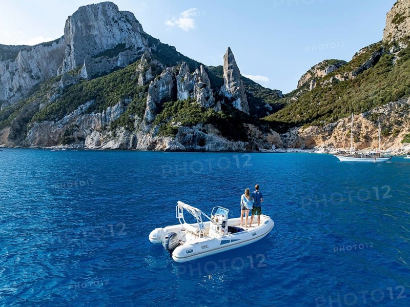 Couple on motorboat off picturesque rocky coast, cliffs with L'Aguglia pinnacle, blue sea and Cala Goloritzé beach, aerial view, Golfo di Orosei, Baunei, Sardinia, Italy