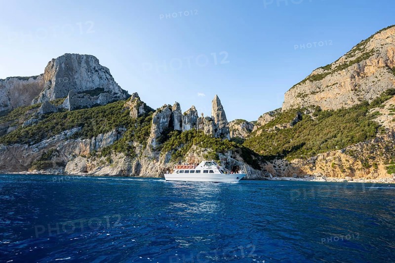 Excursion boat off picturesque rocky coast, cliffs with caves in morning light, L'Aguglia rock pin, blue sea and Cala Goloritzé beach, Golfo di Orosei, Baunei, Sardinia, Italy