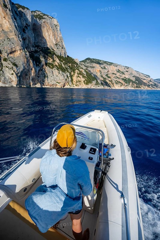 Young woman riding a motorboat along the picturesque rocky coast, cliffs and blue sea, Golfo di Orosei, Baunei, Sardinia, Italy