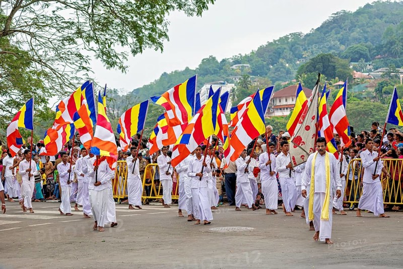 People in white robes carry Buddhist flags at a ceremonial parade, with hills in the background, The Perahera Parade in Kandy Sri Lanka