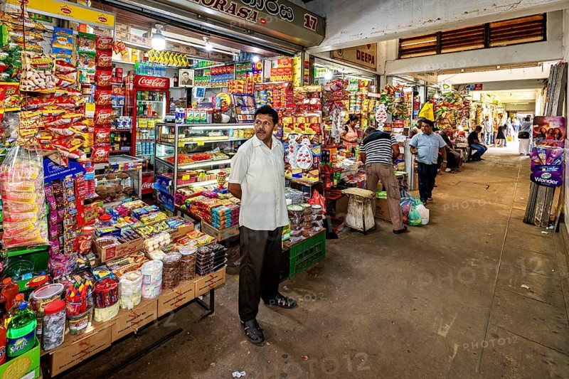 A man stands in a well-stocked shop with numerous colorful foods and drinks, dealers in the market halls of Kandy Sri Lanka