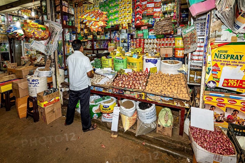 A man in a busy shop surrounded by various colorful packaging and food, dealer in the market halls of Kandy Sri Lanka