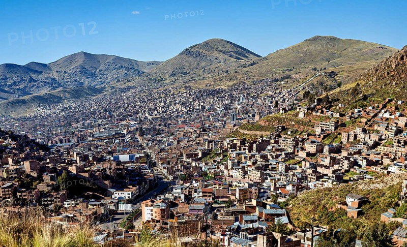 A city nestles on mountain slopes under clear blue skies, lined with green hills and dense buildings in the foreground, a view of the city of Puno in Peru